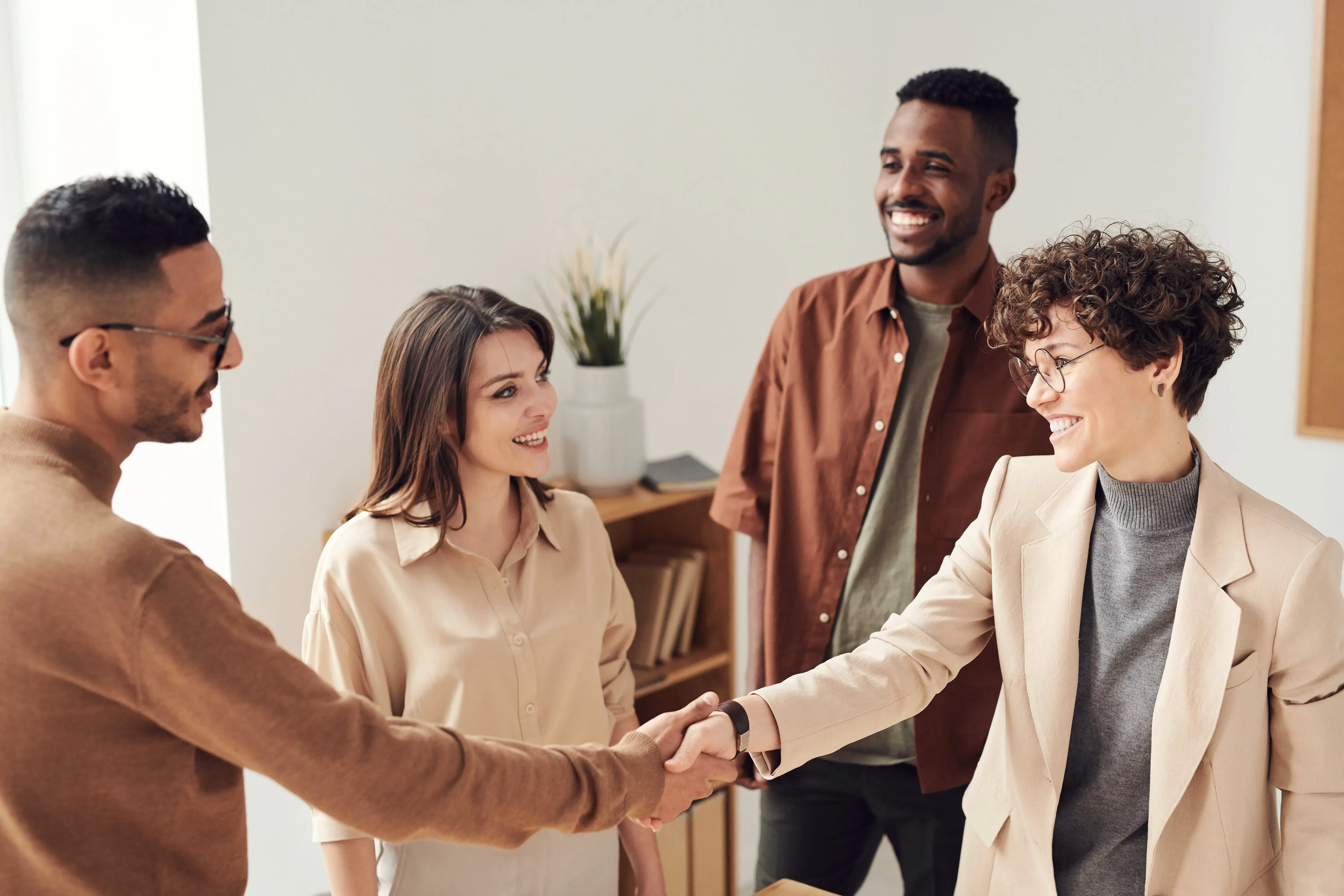Vier lächelnde Personen in legerer Geschäftskleidung, zwei von ihnen schütteln sich in einer Büroumgebung die Hand – ein positives Personalservice-Erlebnis für Bewerber, die nach neuen Karrierechancen suchen.