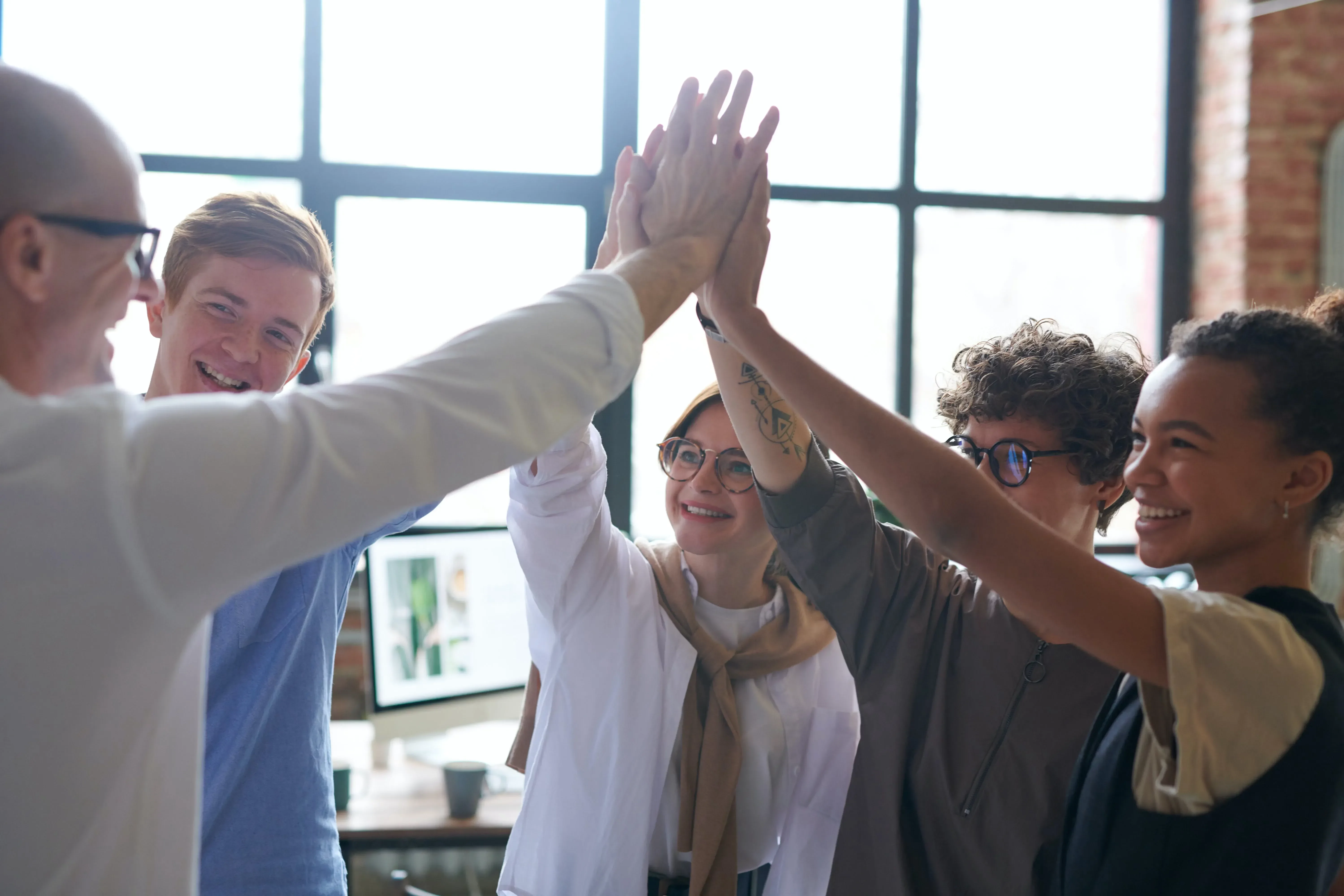 Ein fröhliches fünfköpfiges Team feiert Erfolge mit einem gemeinsamen High-Five in einem hellen Büroraum und spiegelt damit ihren ausgeprägten Sinn für Teamarbeit und gemeinsame Karriereziele wider.